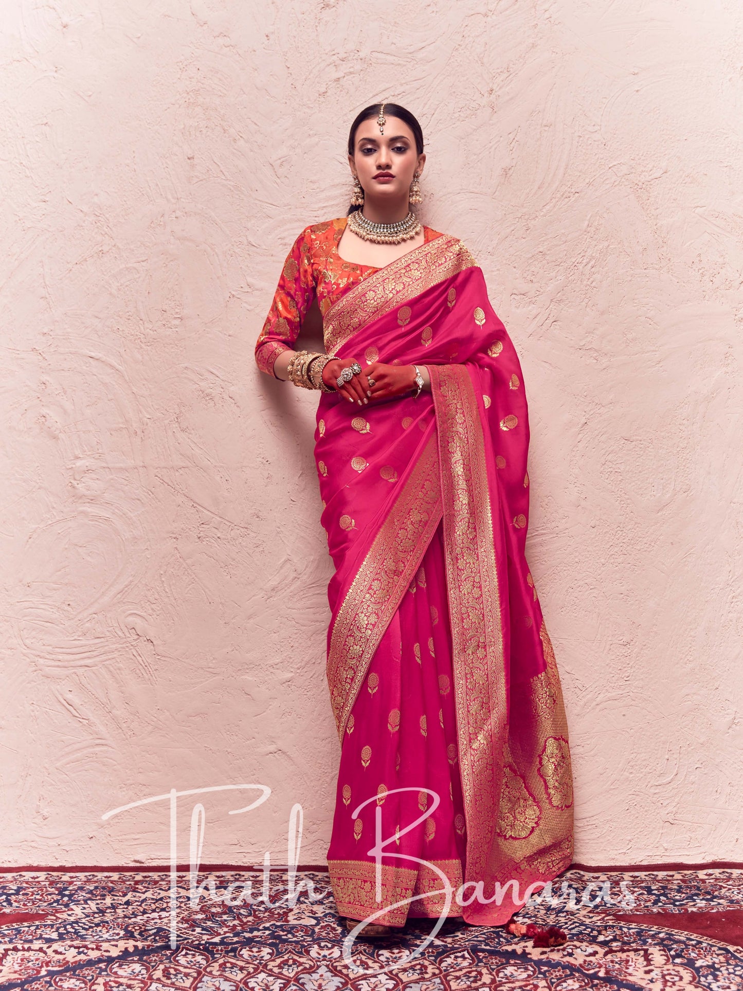 A woman posing in a traditional magenta pink saree with zari weaving and a matching blouse, against a neutral background.